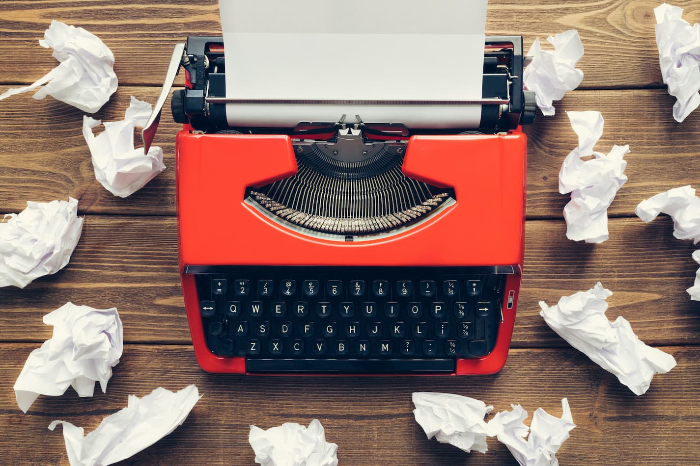 An image of a red typewriter surrounded by crumpled paper on a wooden slat background.