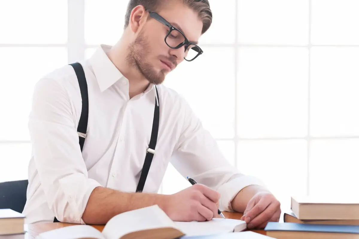 An image of a young man in a shirt and suspenders writing something on a notepad while sitting at his working place.