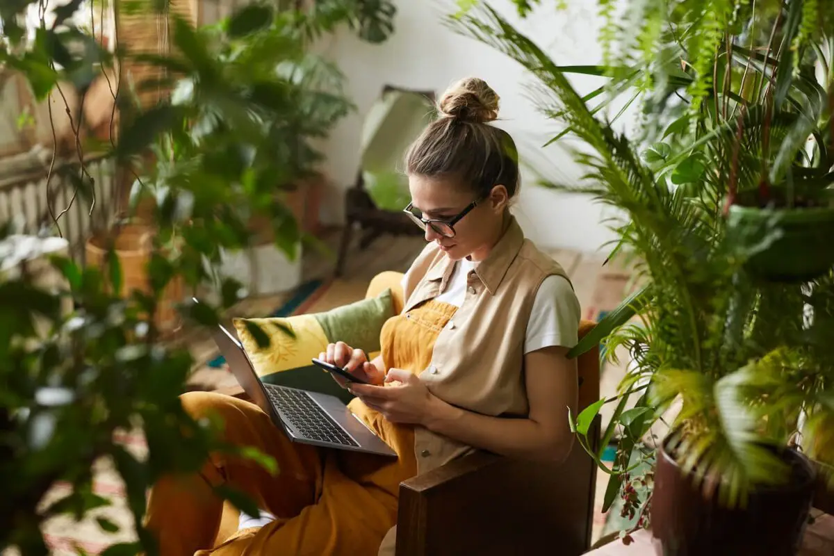An image of a woman working on her laptop and phone surrounded by plants.